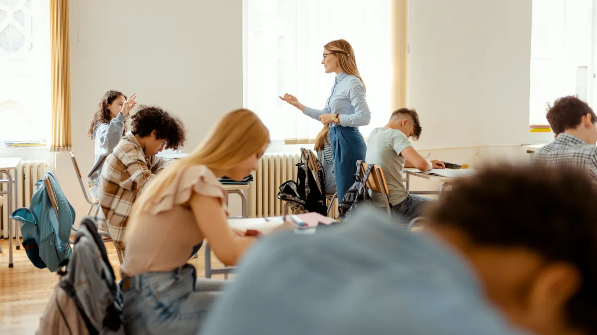 An adult stands in a classroom with students seated at desks. Some students have their hands raised.