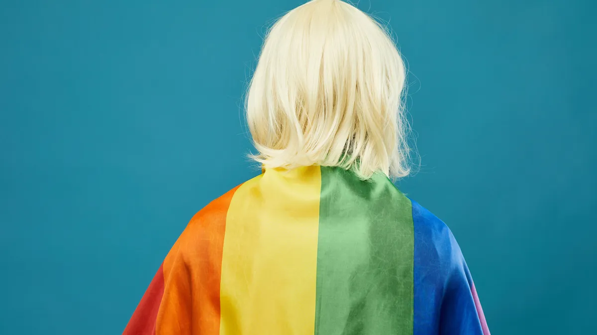 A person with a shoulder-length blonde wig drapes themselves in a rainbow flag.