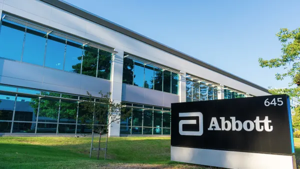 Modern glass and metal office building with a reflective blue-tinted facade, set against a clear blue sky. In the foreground, a large black and white sign reads “Abbott” with the company’s stylized logo and the number “645.” The area has neatly cut grass and a few small trees.