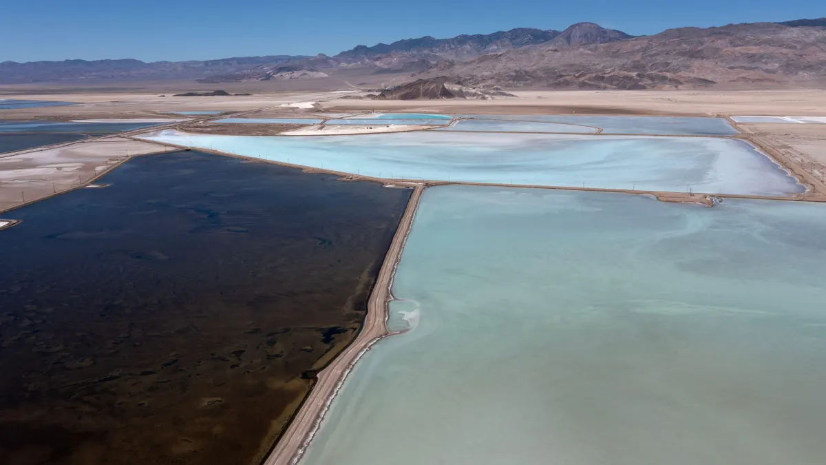 Aerial view of the Silver Peak Lithium Mine, showcasing a vast desert landscape with a network of rectangular evaporation ponds in varying shades of blue and green. These geometric pools are interlaced with earthen pathways and surrounded by arid terrain, illustrating the industrial scale of lithium extraction in a remote environment.