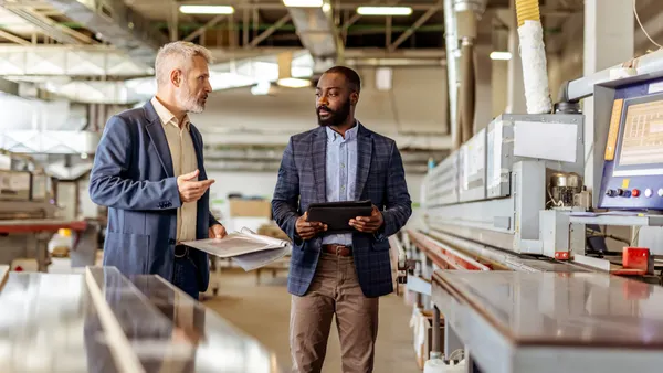Two professionals interact in a manufacturing facility, discussing operations and reviewing data using modern equipment.