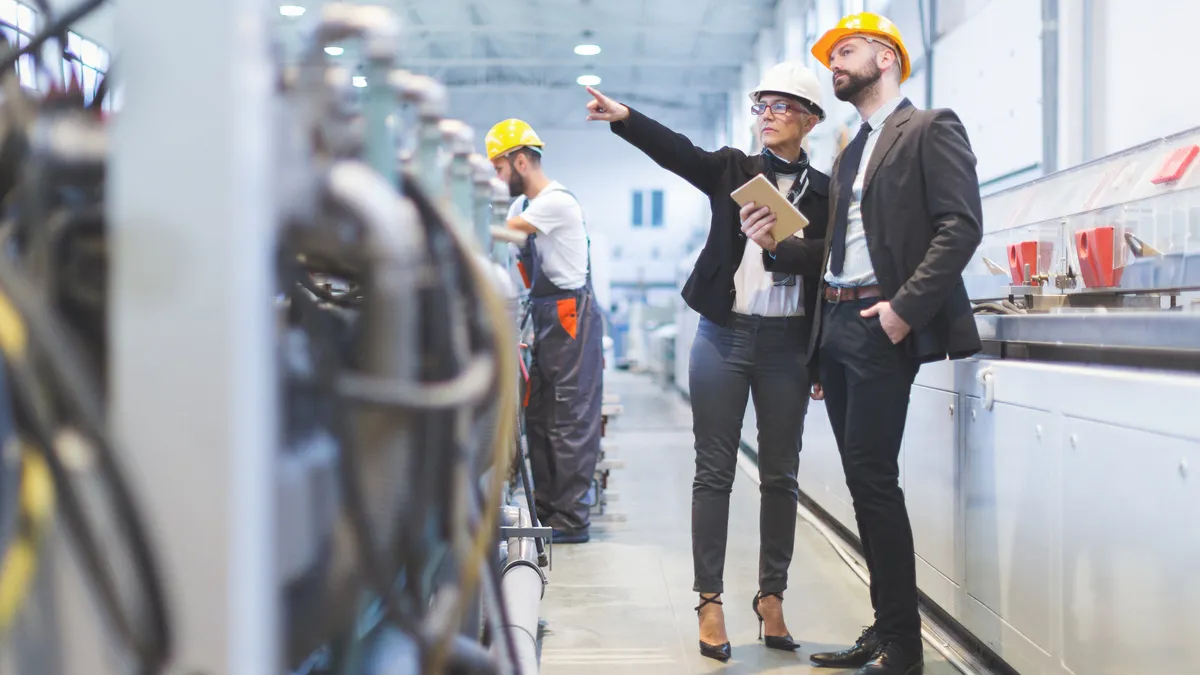 Two people dressed in business clothes stand in the foreground of a factory setting. The person on the left is pointing at something, the person on the right following along.