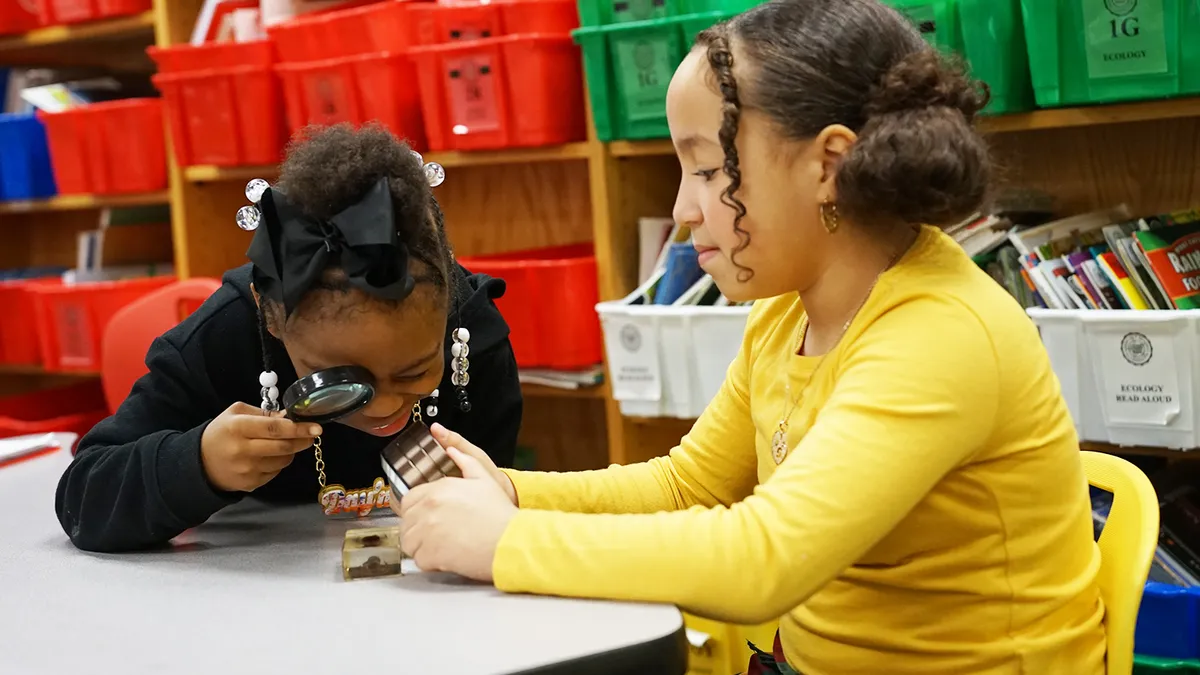 Two students sit at a table with shelves behind them. The two students are looking at an object on the table with one student holding a magnifying glass at the object.