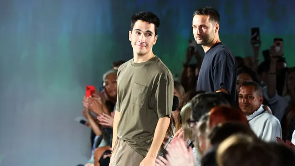 A side view as two men in T-shirts walk down a runway. One turns toward the crowd on the sidelines and smiles.