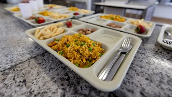 Two rows of food trays lay side by side on a table in a school cafeteria.