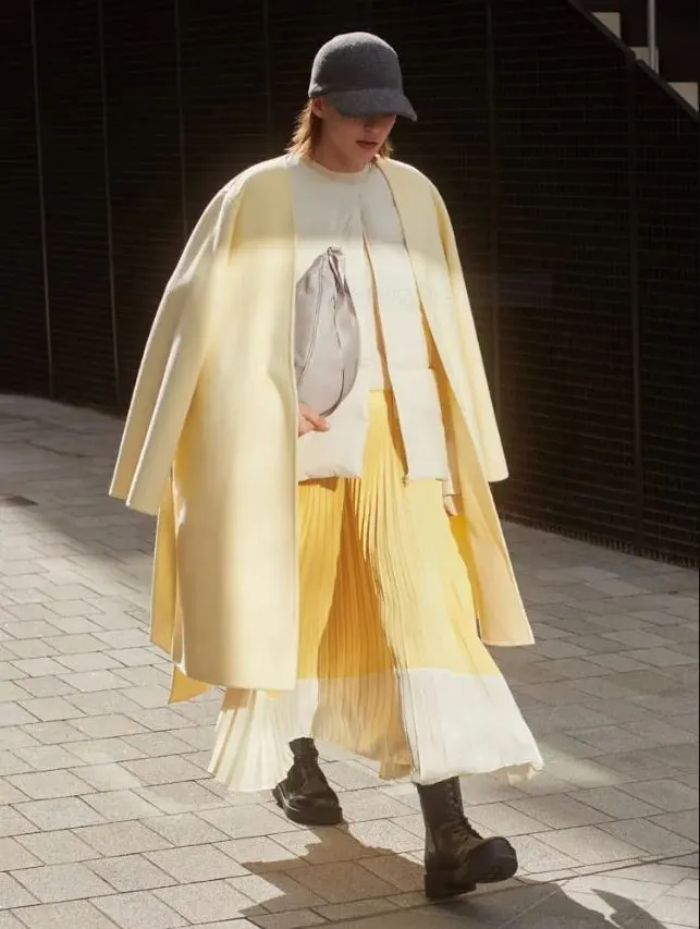 A person wearing a yellow coat and flowing outfit from the Uniqlo collaboration with Clare Waight Keller walks down the street.