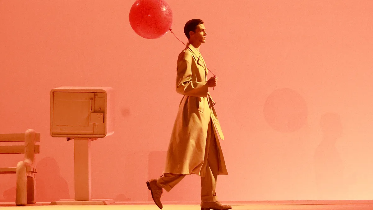 A person walks across a stage holding a red balloon and wearing a trench coat.