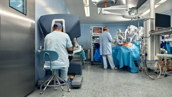 A clinical operating room with two medical professionals; one seated at a robotic surgery console and the other standing next to a patient covered in blue drapes with robotic arms positioned above.