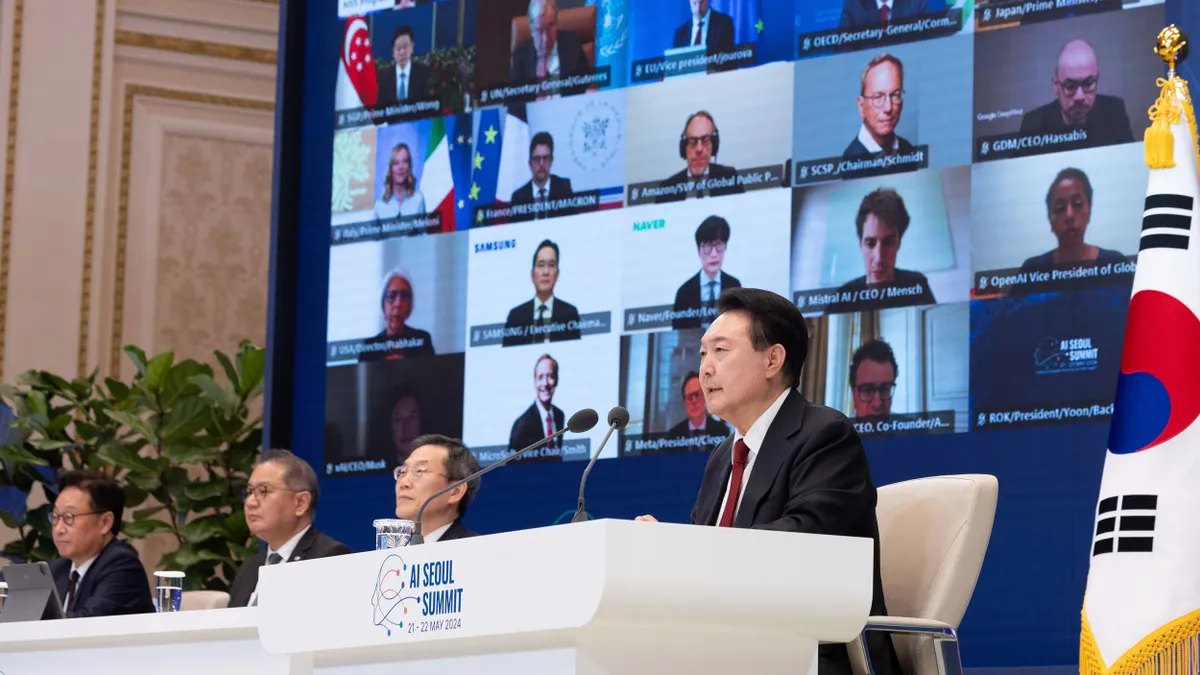 South Korea's president speaks with several executive dressed people, a large screen behind the people projects multiple people on a virtual call.