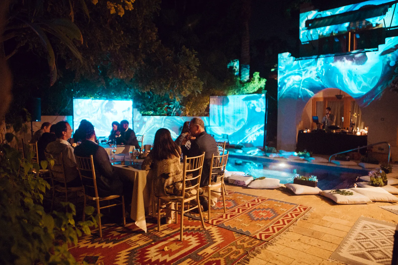 People sit around outdoor tables on top of woven rugs with blue light patterns projected on walls and screens and tropical foliage to the left. A pool and people preparing food are in the background.