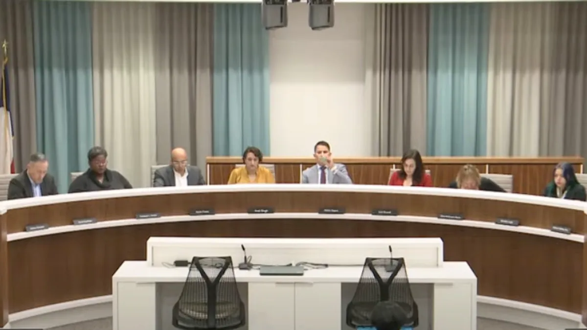 A panel of 10 adults sit at a desk on a dias. Behind them are curtains and flags.