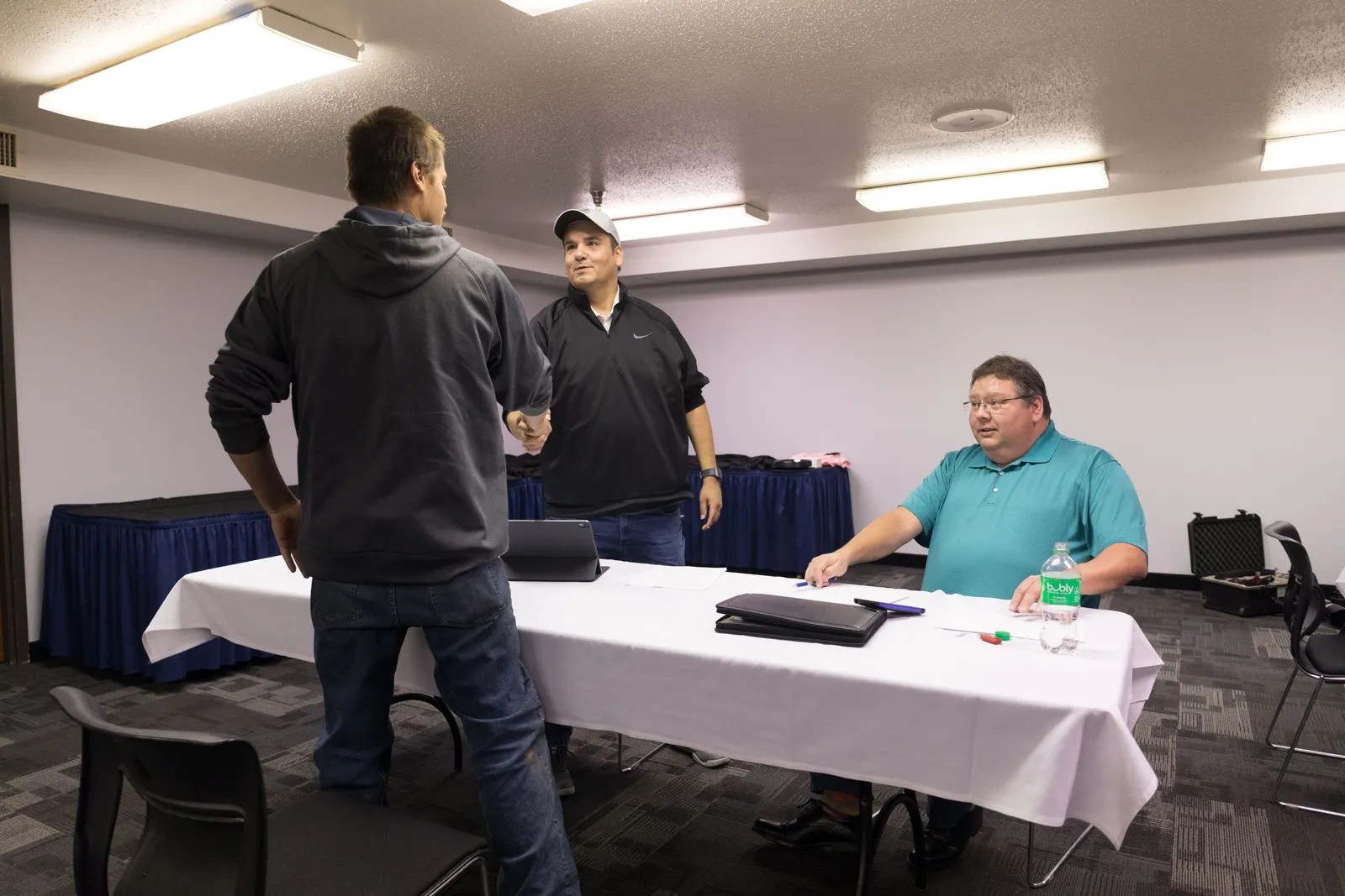 A person shaking hands with two men seated behind a table.