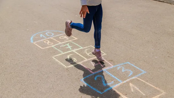 A student is shown below the shoulders skipping across numbers chalked in asphalt on the ground. The students' shadow is also seen.