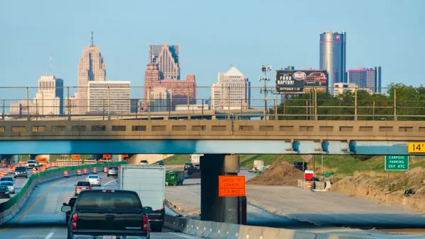 Cars and a truck on the interstate near Detroit, driving below an underpass with the cityscape in the background.