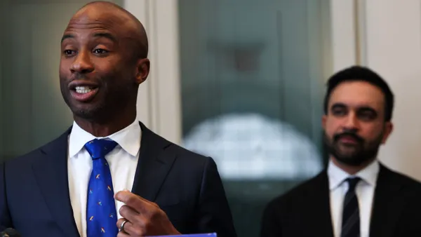 Kamar Samuels speaks at a podium as Zohran Mamdani listens behind him.