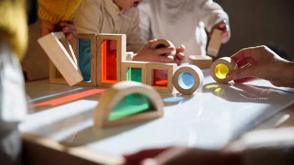 Small children are playing with wooden blocks with colorful see-through windows. An adult hand is seen in the photo