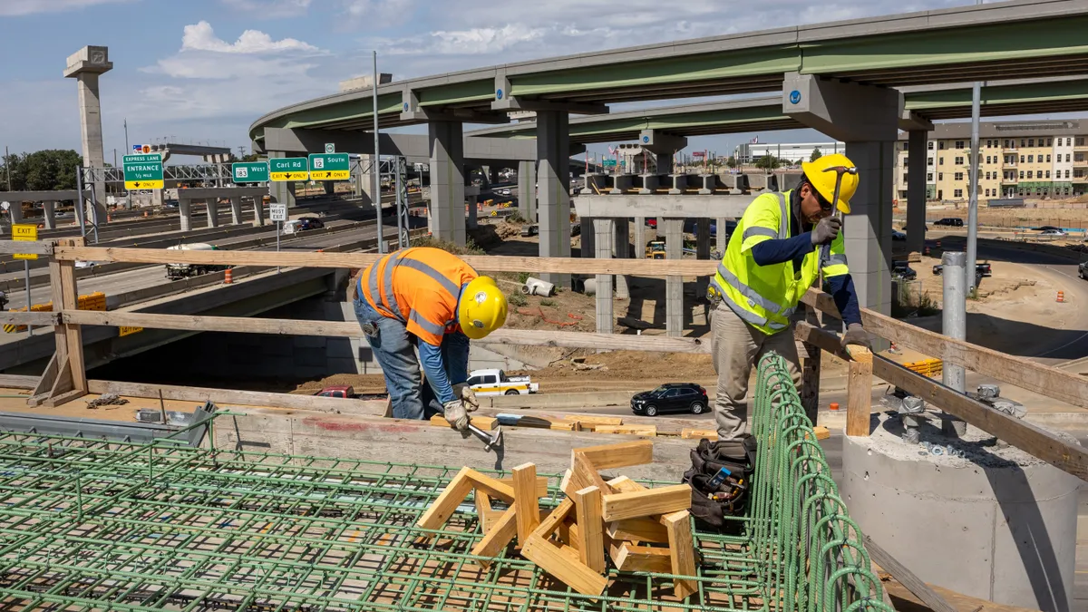 Two construction workers in reflective vests and hard hats hammer a wooden frame with a highway overpass in the background.