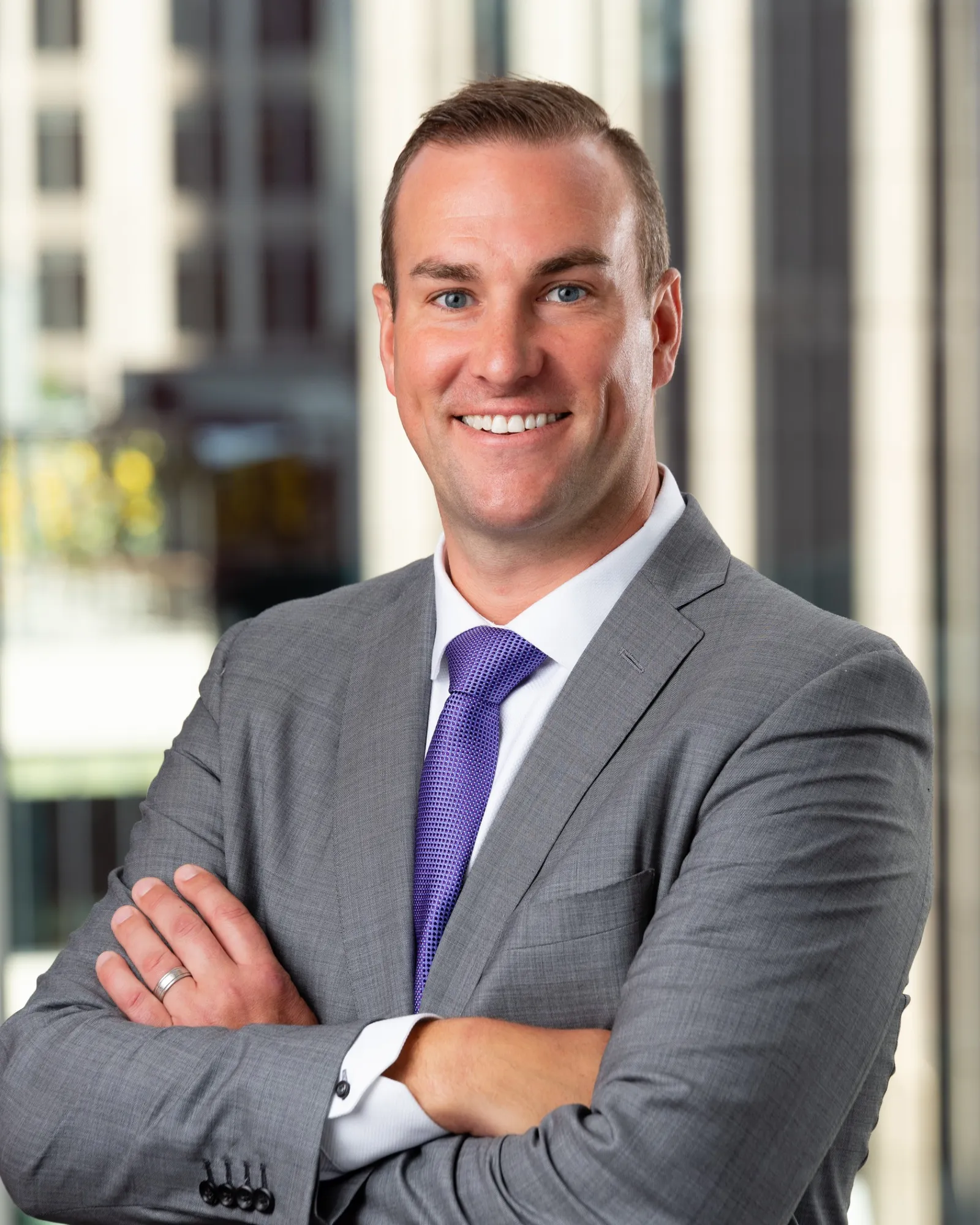 A person wearing a suit and tie stands with their arms crossed and smiles.