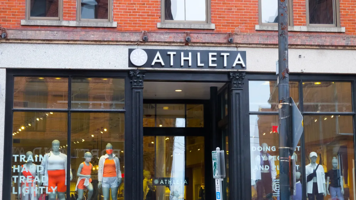 Mannequins in orange and white stand in a store window to left of a black-trimmed doorway of an Athleta store, others in black and white are on the right.