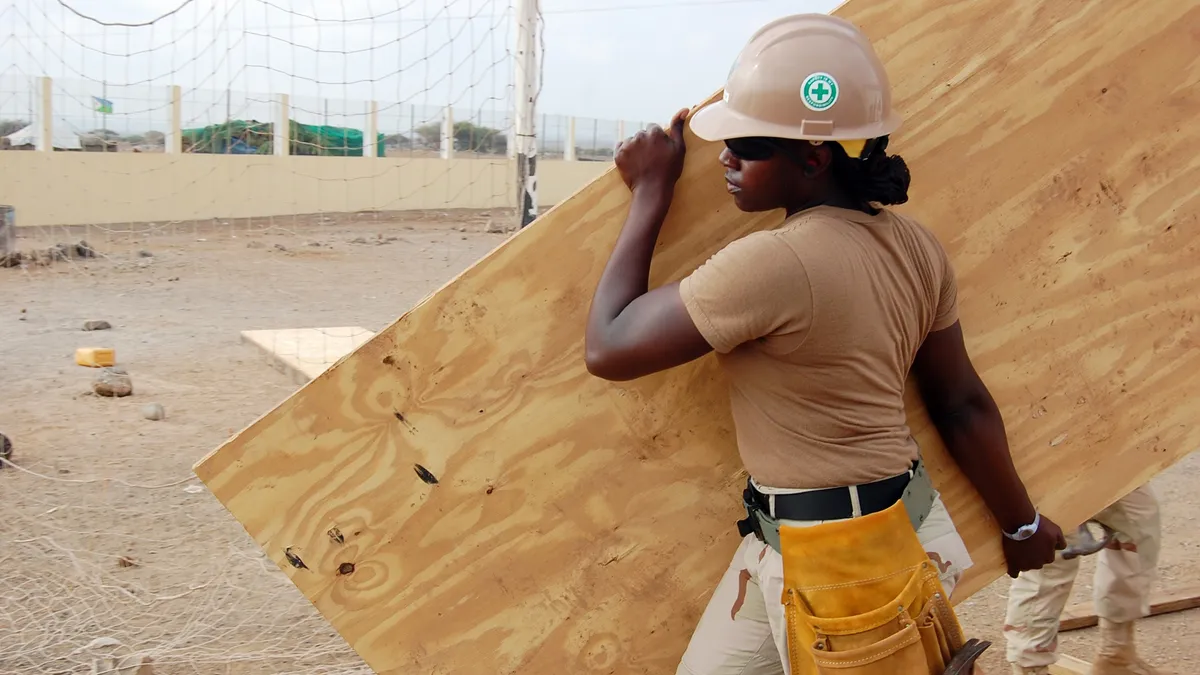 A Black woman in a yellow hard hat carries a large piece of plywood.