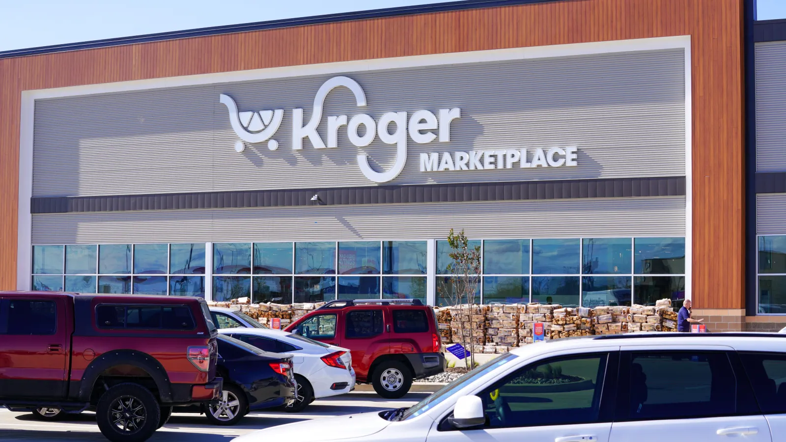 Exterior of a supermarket with a white sign reading "Kroger Marketplace" and cars parked in front.