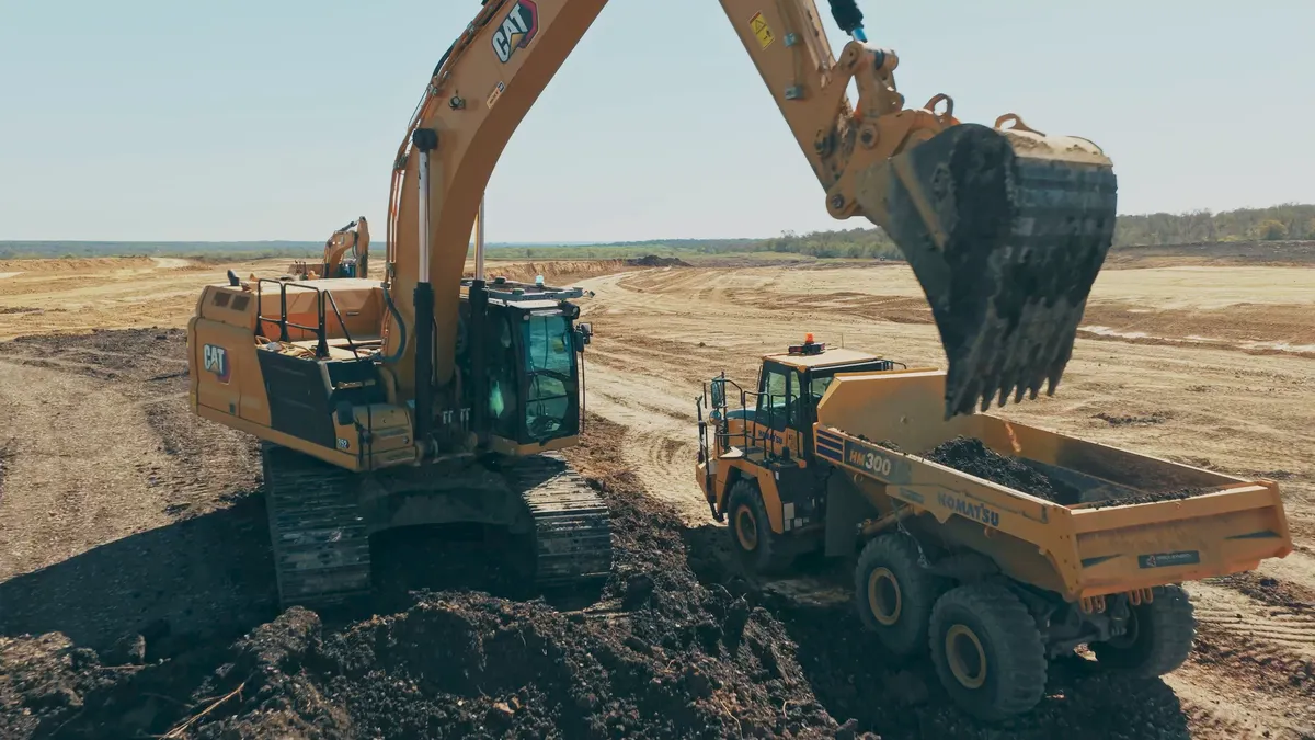 A piece of heavy machinery near another piece of machinery. Both are yellow and are framed under a blue sky on a plot of dirt.