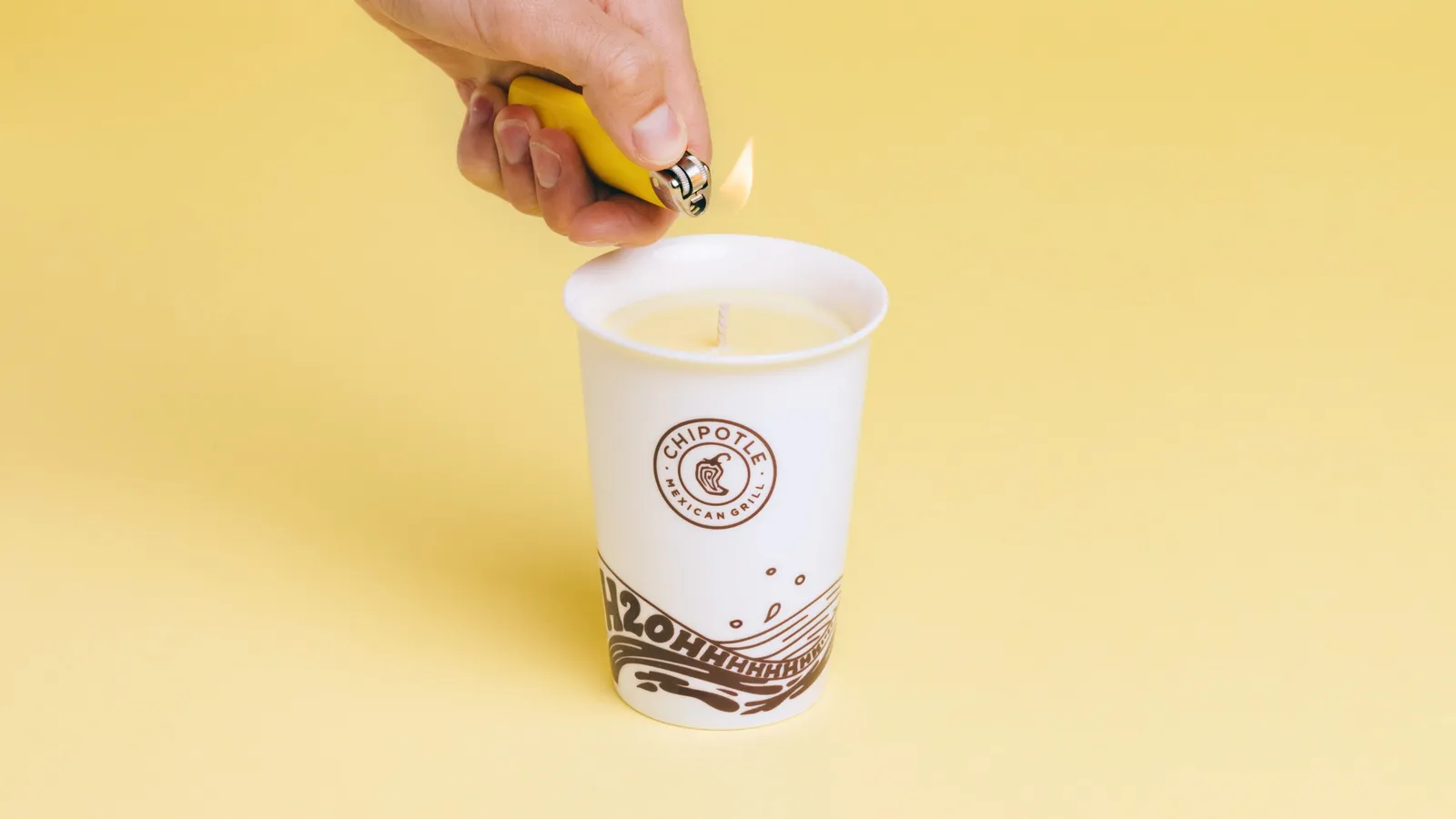 A hand lighting the white and black Chipotle water cup with a yellow candle inside.