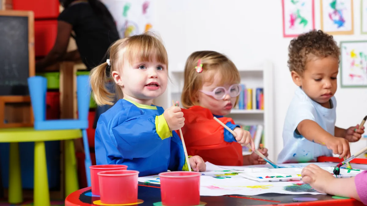 Three young children are sitting at a desk. They are wearing aprons and have paint and paper on the desk. They are holding paint brushes.