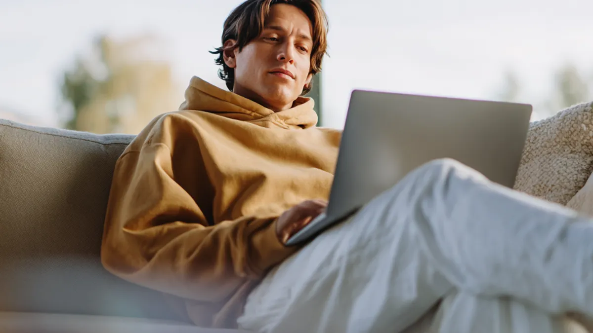 A person in a tan hoodie sits on a couch using a laptop, with natural light coming from a window behind them.