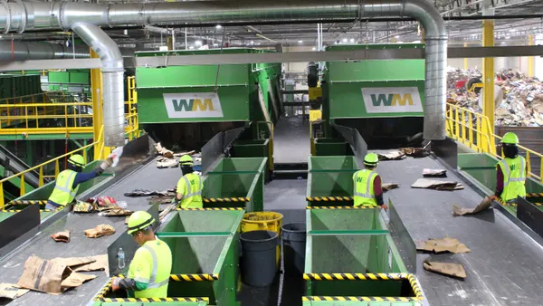 Six workers wearing bright yellow vests with WM's logo work next to conveyor belts at a recycling center.
