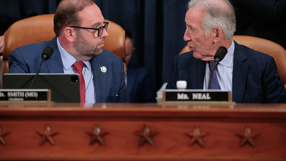 Two people are seated at a table with microphones in a room. They are looking at each other