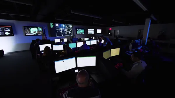 People sit at desks, in front of illuminated computer monitors, in a dark room that has large computer screens on the back wall
