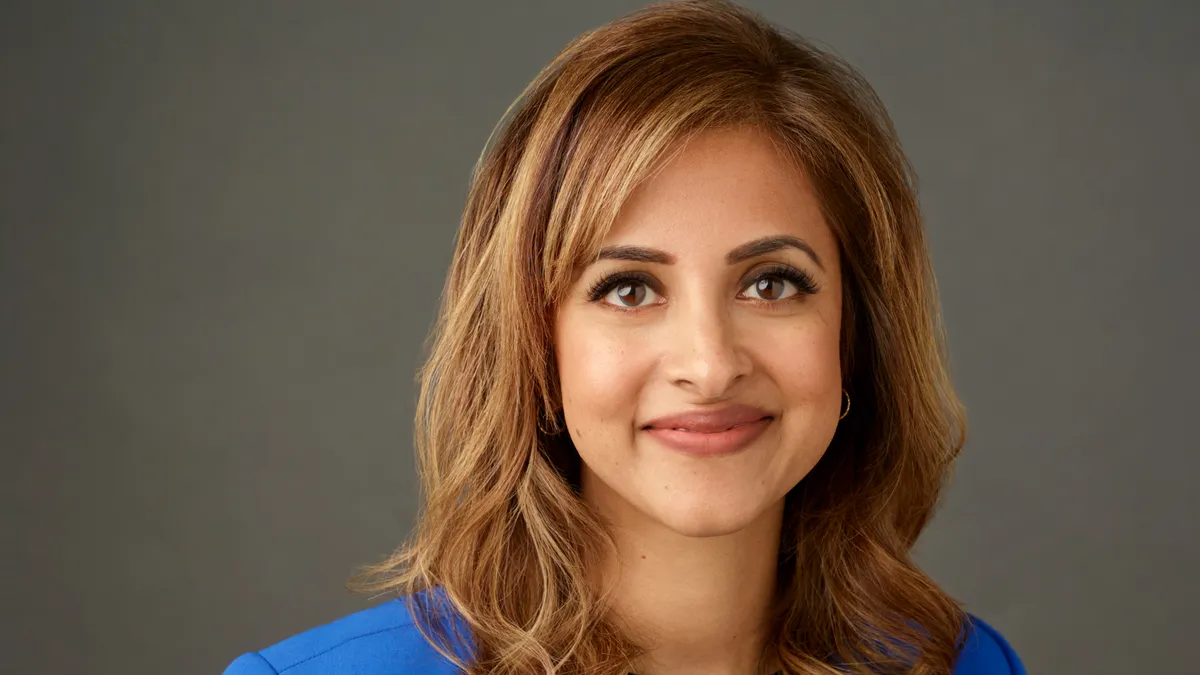 A smiling person with long brown hair, wears a bright blue jacket.