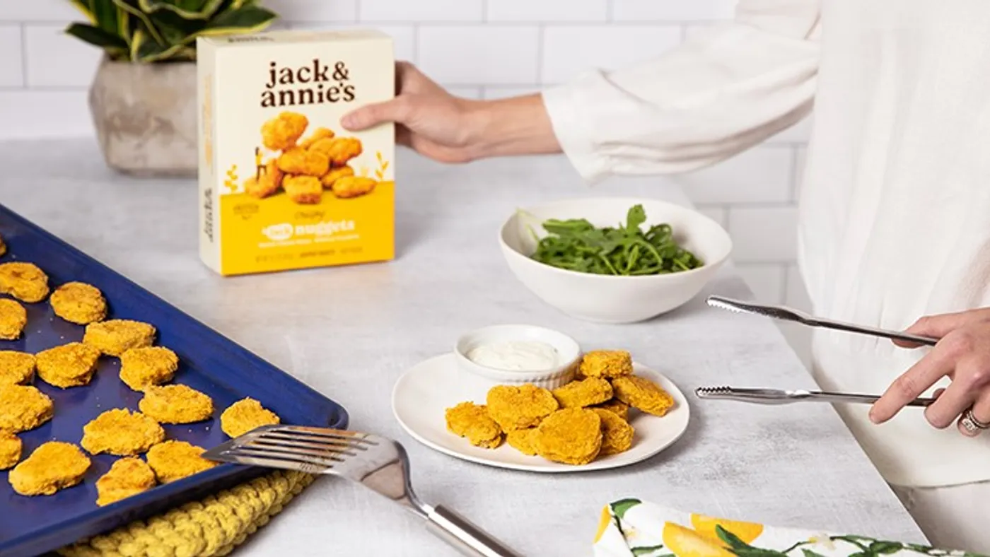 A person stands next to a white kitchen counter holding a box of Jack & Annie's nuggets. There is a baking tray full of nuggets and a plate with nuggets and white dip on the counter in front.