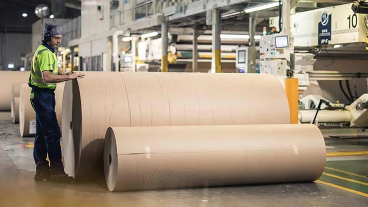 A person wearing protective equipments stands next to a several large paper rolls in a factory.