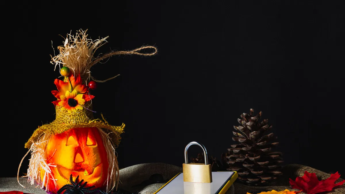 Cell phone with a yellow case, with the screen on and a locked padlock on top next to a Halloween pumpkin adorned on sackcloth and surrounded by toy spiders and fallen leaves next to pine cones.