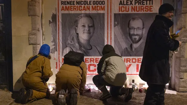 Three people kneel and one person stands in front of large signs that say "Renee Good, American Mom" and "Alex Pretti, ICU Nurse"