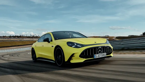 A yellow vehicle drives on a test track, with rolling hills and clouds in the background.