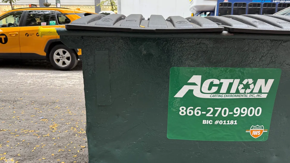 A commercial dumpster sits on the sidewalk next to a busy avenue with a taxi. The dumpster bears the "Action Environmental" logo.
