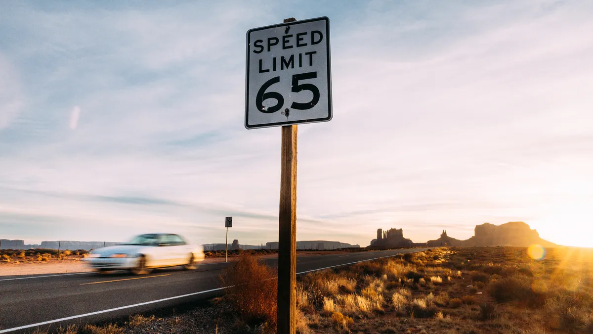 A car passes a sign reading "Speed Limit 65" on a desert highway.