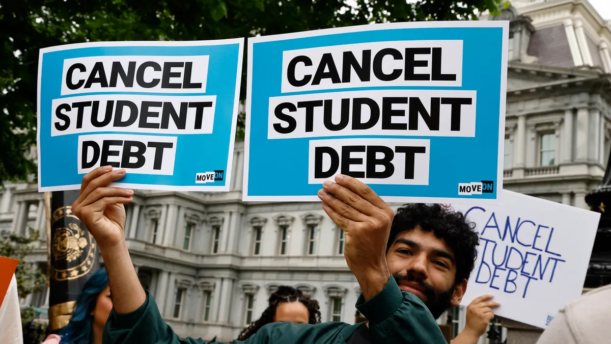 At a rally, a man hold up two small signs, one in each hand. Both signs say "cancel student debt."