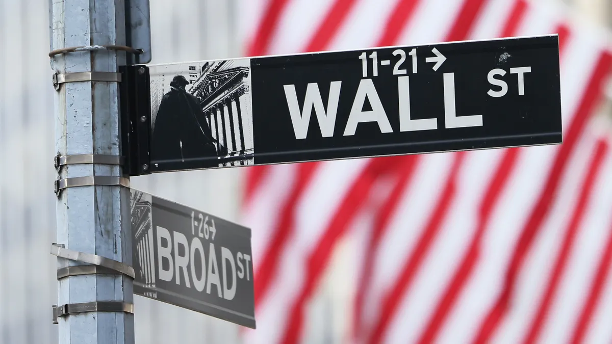 Street signs for the intersection of Wall St. and Broad St., with the stripes of an American flag in the background.