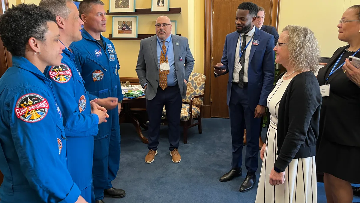 A group of adults stand in a room facing each other. Several of the adult have blue uniforms.