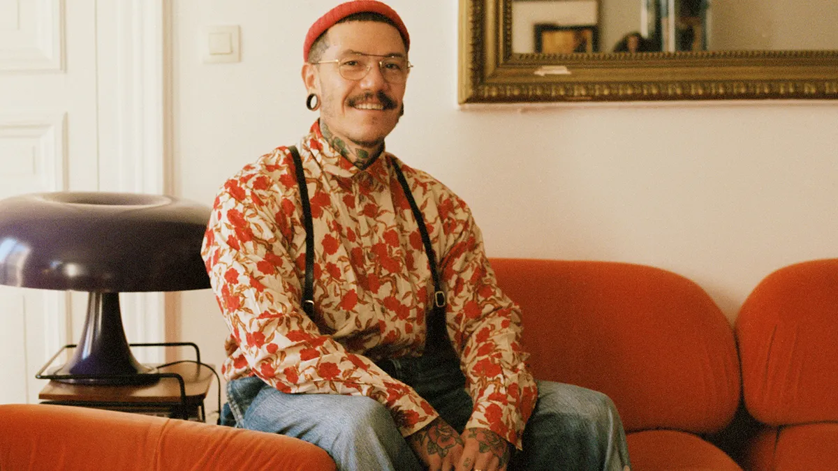 A person with glasses, a mustache, and tattoos on his neck and face wears colorful clothing and sits smiling at the camera.