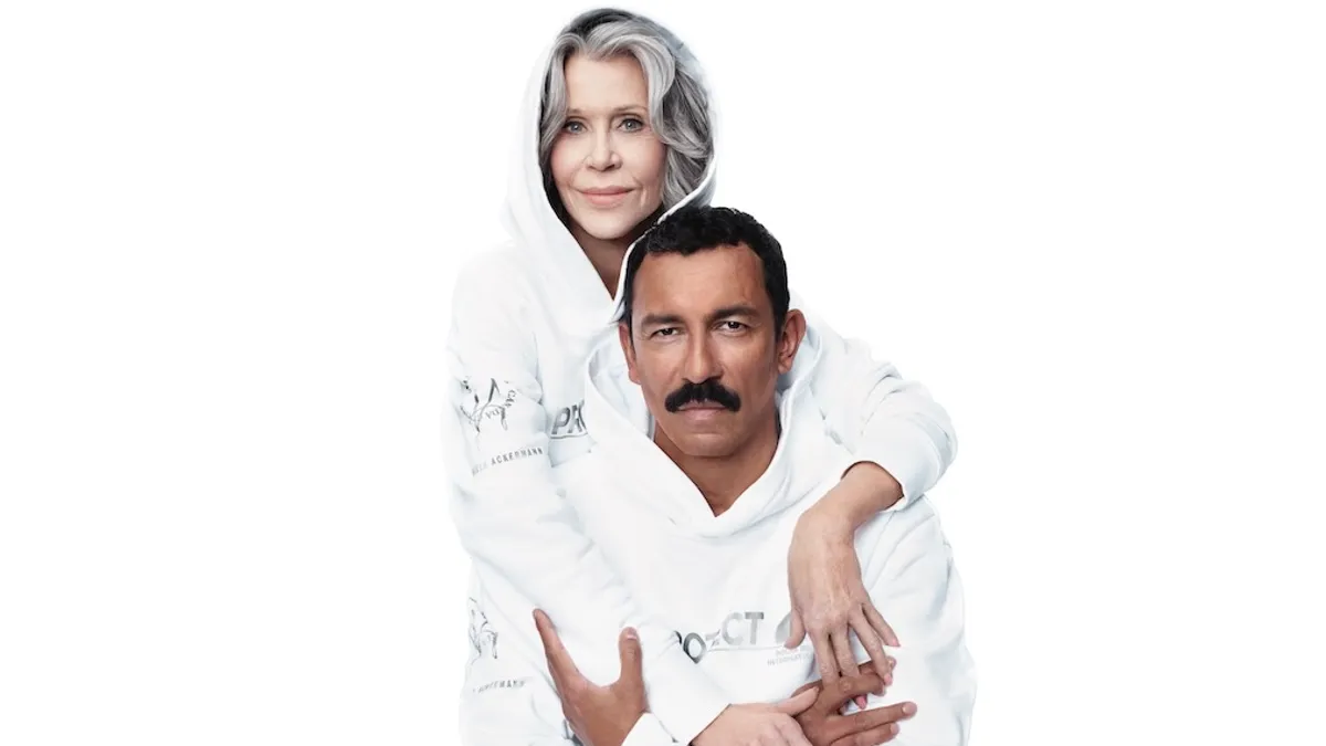 Two people, both wearing white, one in a hoodie, the other a mustache, embrace.