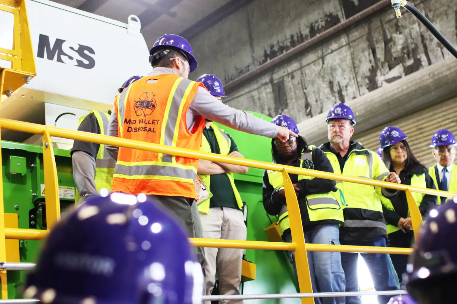 A worker gives a tour at a MRF to people wearing hard hats and safety vests.