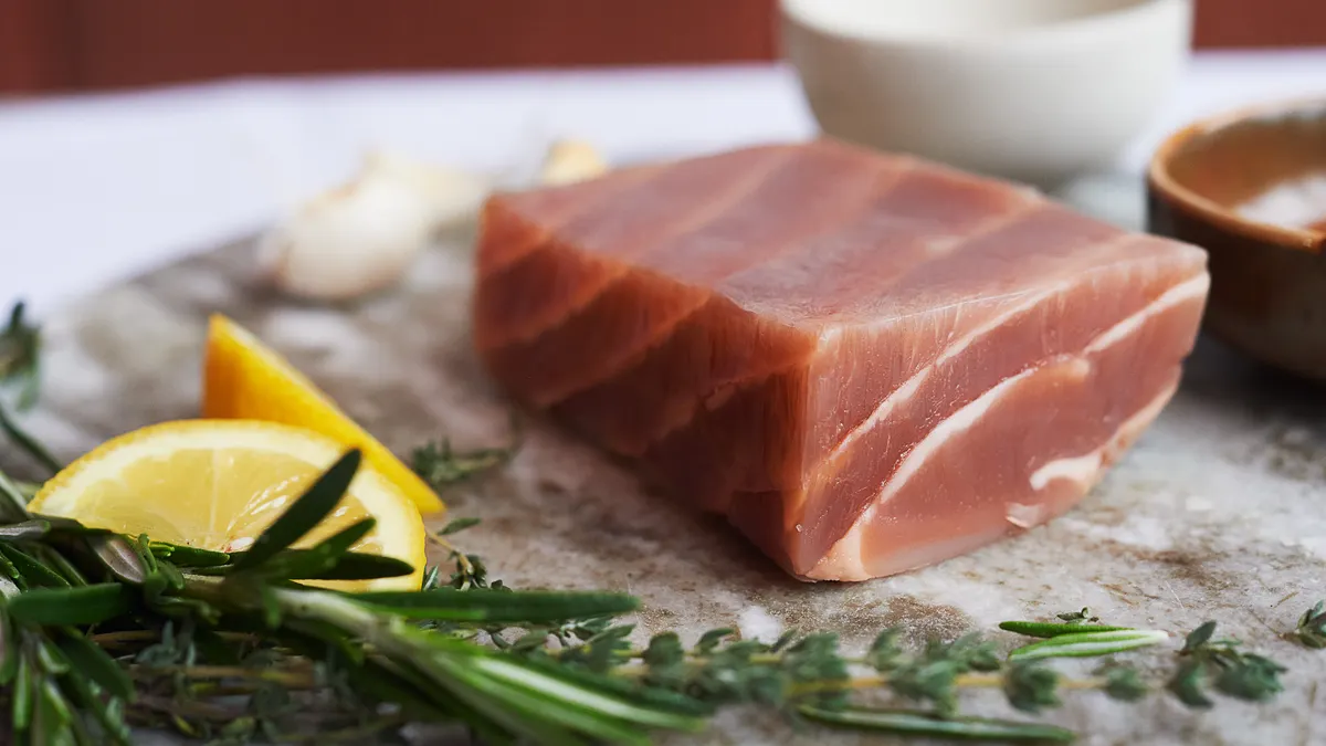 A raw plant-based salmon filet made by New School Foods on a marble surface with lemon slices and herbs in the foreground and bowls in the background.