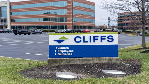 A white, blue and green Cleveland-Ciffs sign and a parking lot and a red-bricked building with windows in the background.