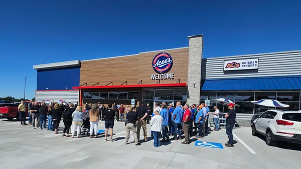 A photo of the ribbon cutting at a travel center. A number of people stand outside on a clear day next to a building. Signs on the building say Acee's. Community Driven. AC's chicken fingers."