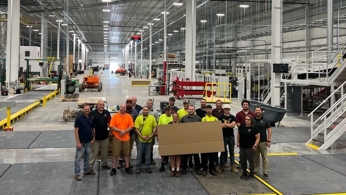 A group of workers hold up a sheet of corrugated medium in an industrial facility.